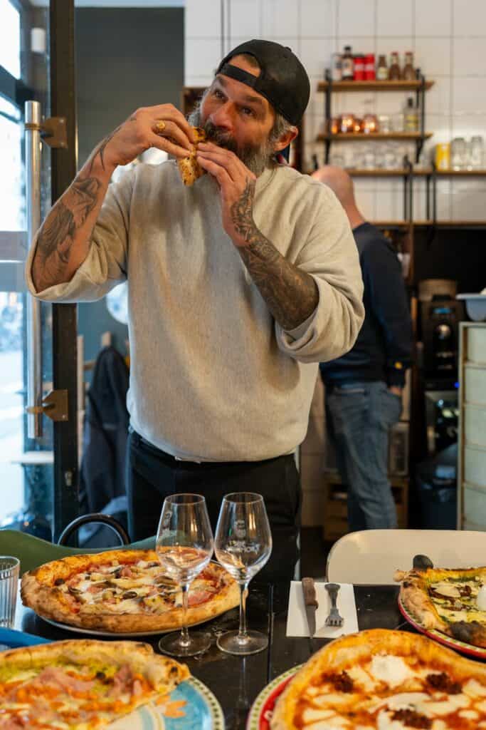 Pépé Ronchon dégustant une part de pizza devant une table garnie de plusieurs pizzas.