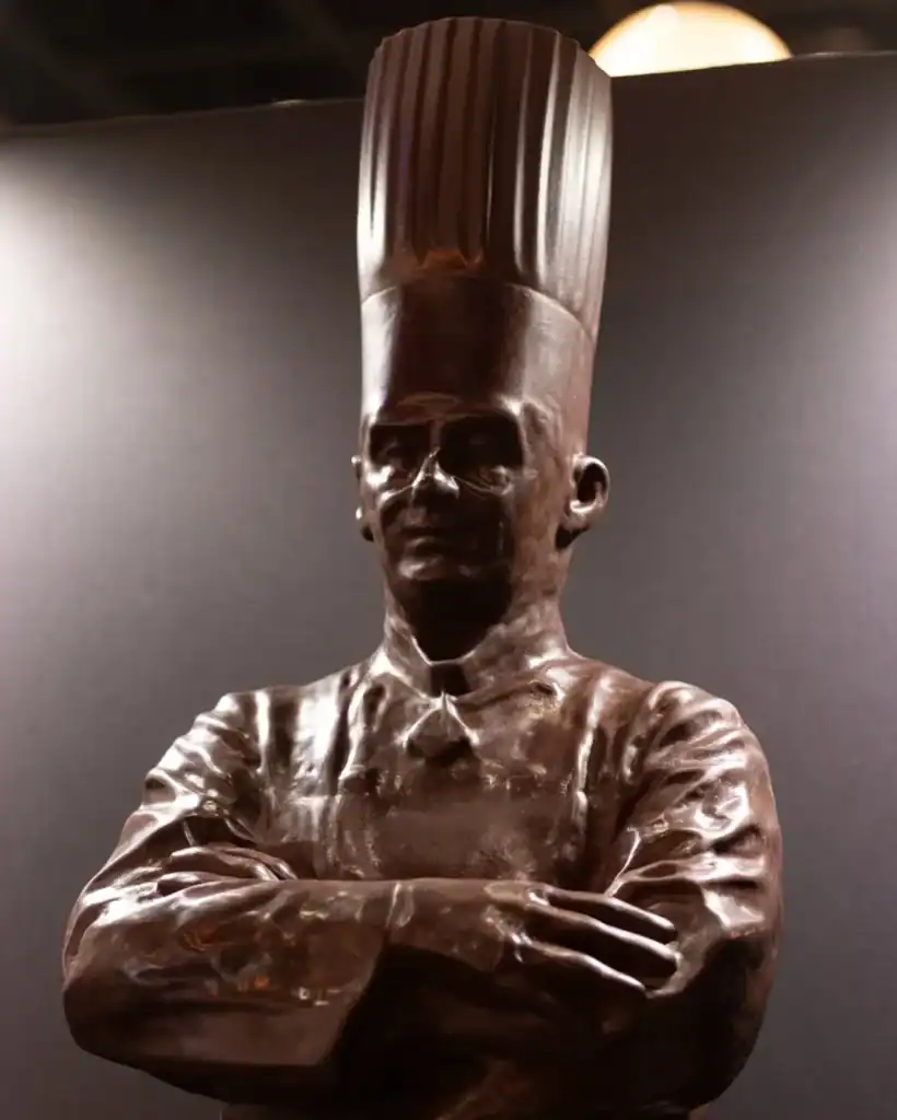 Statue de Paul Bocuse vue de face, coiffé de sa haute toque et les bras croisés sur sa veste de cuisinier.